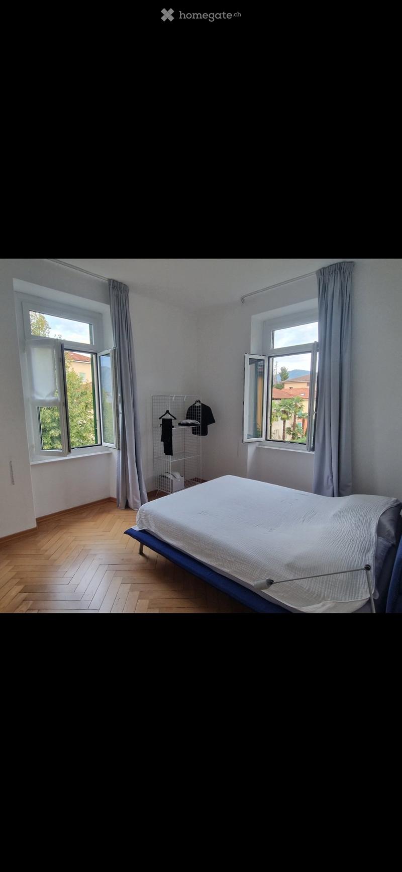 Bright bedroom with wooden flooring, large windows with open blinds, a neatly made bed, and a small clothing rack with hanging items.