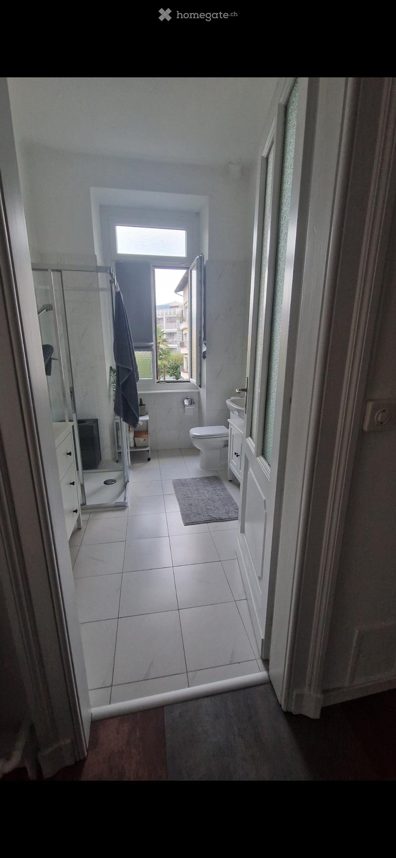Modern bathroom with white tiles, a toilet, sink, mirror, and a small shower stall. A towel hangs on a rack, and a small rug is placed on the floor.