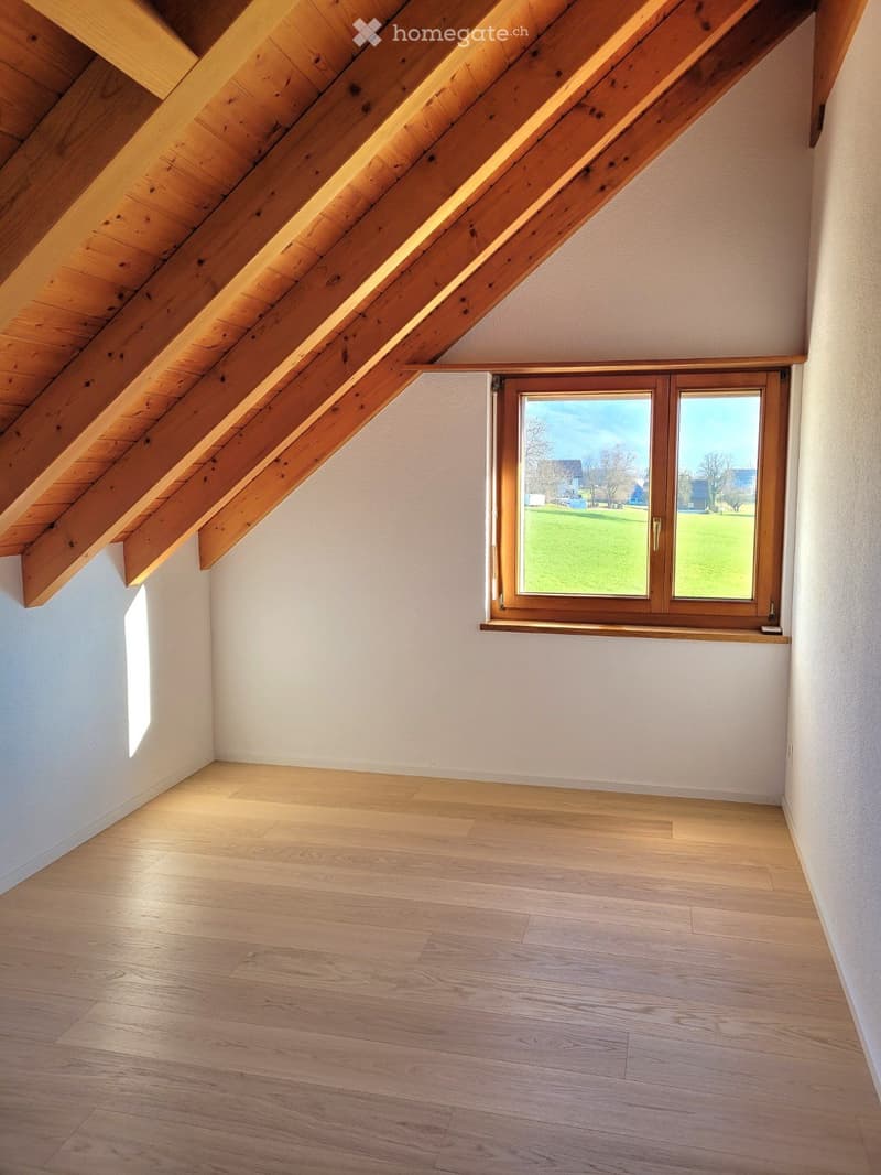 An empty room with wooden flooring, white walls, and wooden beam ceiling, featuring a window with a view of a grassy field.