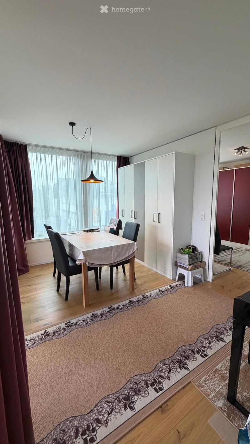 Dining room with a wooden floor, large window with curtains, dining table with chairs, cabinets, and a patterned rug.