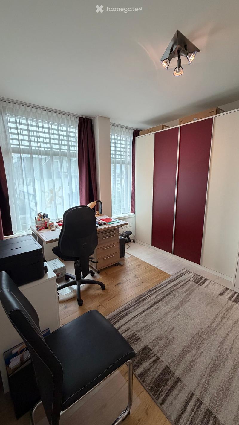 A furnished room with a wooden floor, large windows with curtains, a desk with a chair, a printer, and a cabinet.