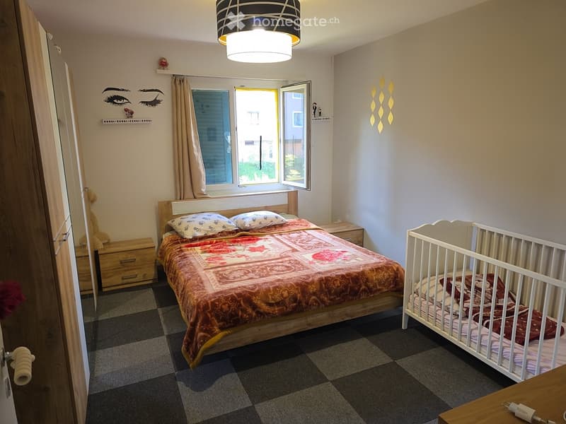 Cozy bedroom with a double bed, crib, nightstand, and decorative wall art. Features a large window with curtains and a modern pendant light.