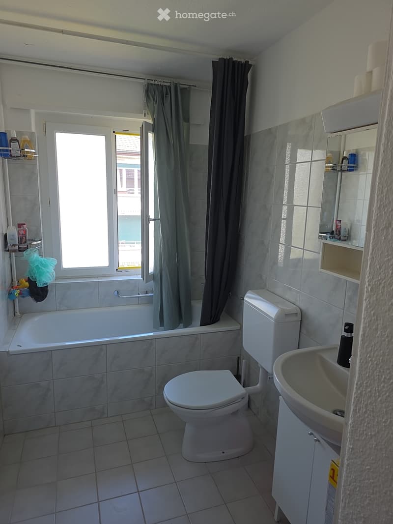 Bathroom with a bathtub, toilet, sink, mirror, and shelves. The room has tiled walls and a window with a curtain.