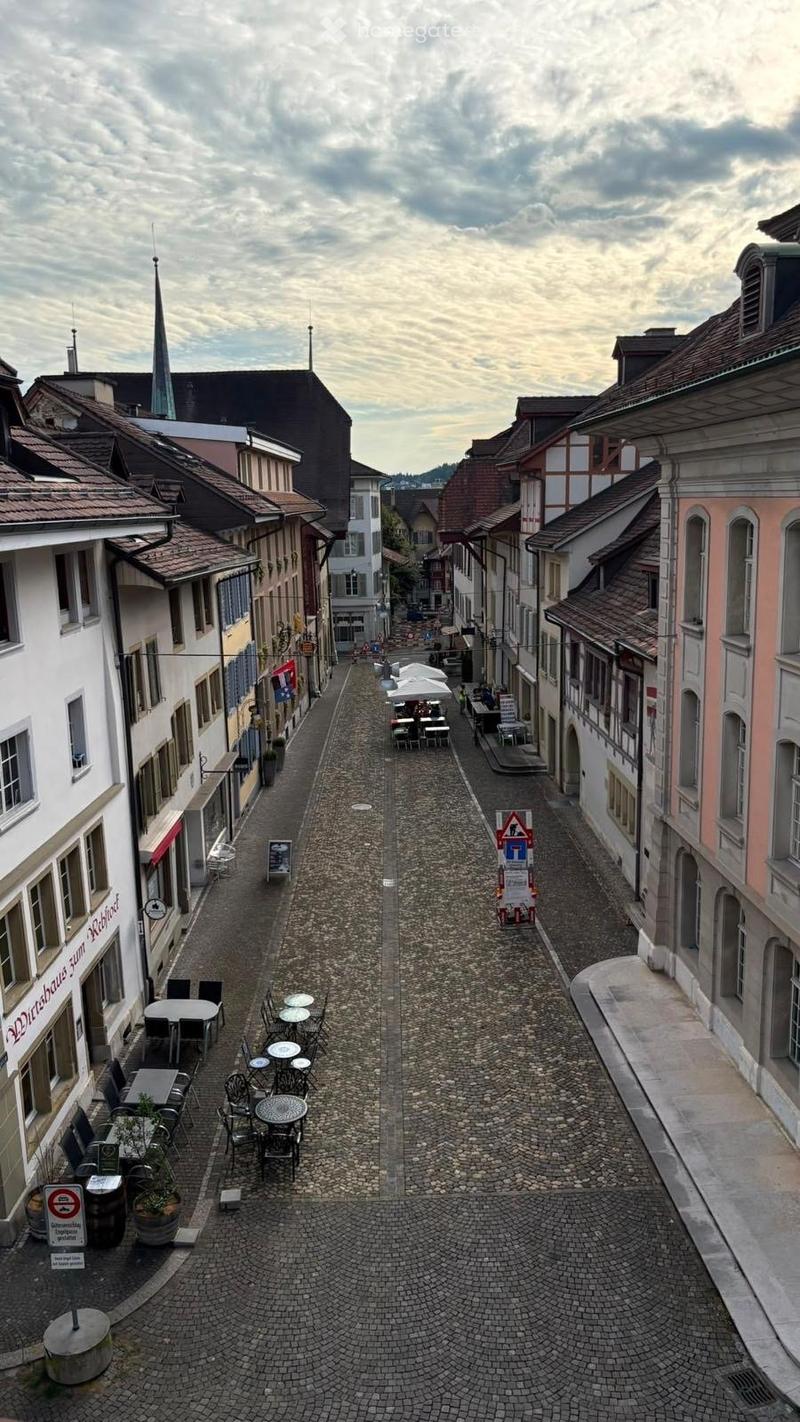 A narrow cobblestone street lined with historic buildings, featuring outdoor seating areas with tables and chairs, and a few pedestrians visible in the distance.