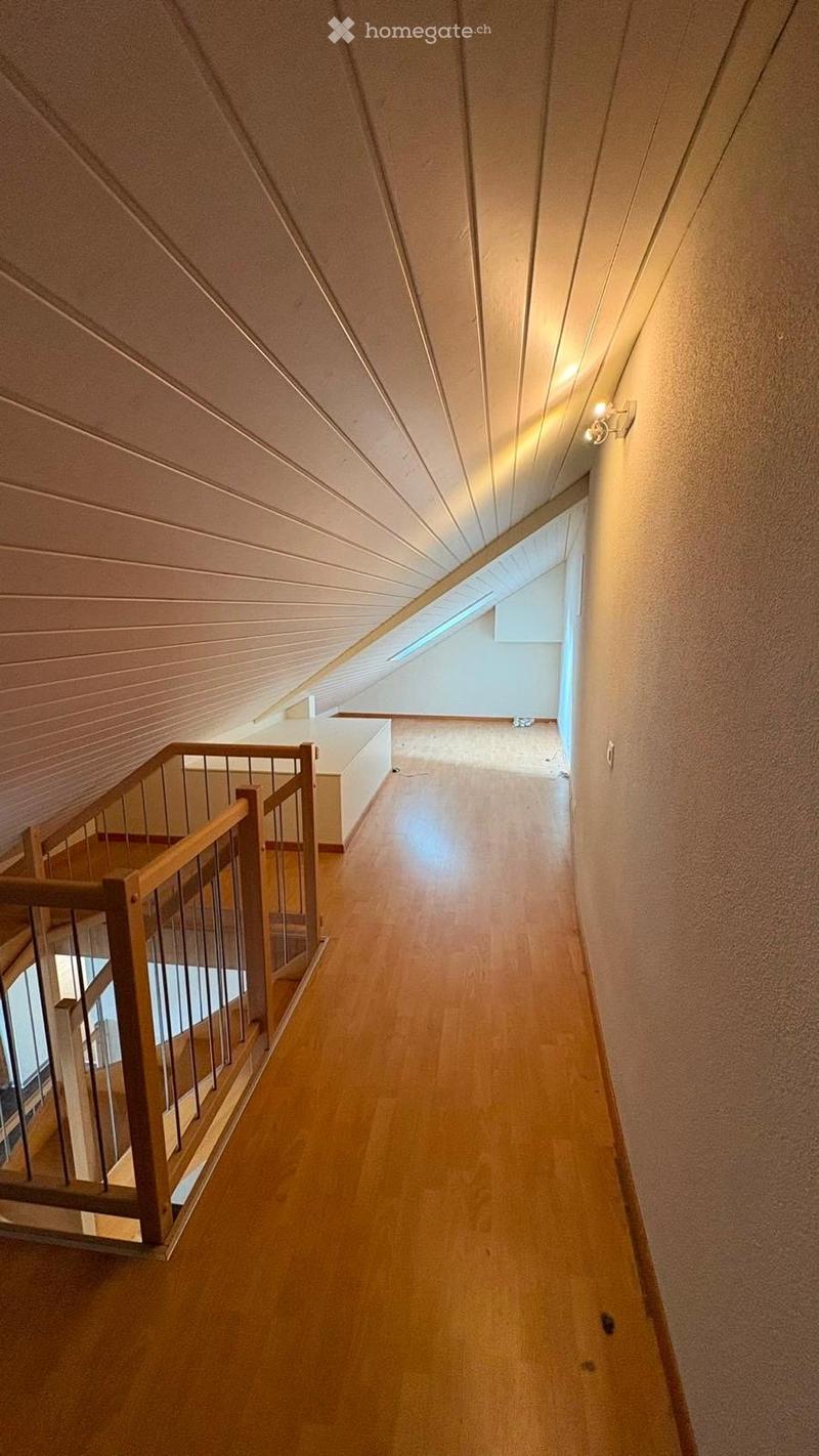 A narrow hallway with wooden flooring, slanted ceiling, and a wooden railing on one side. Natural light filters in from a skylight at the far end.