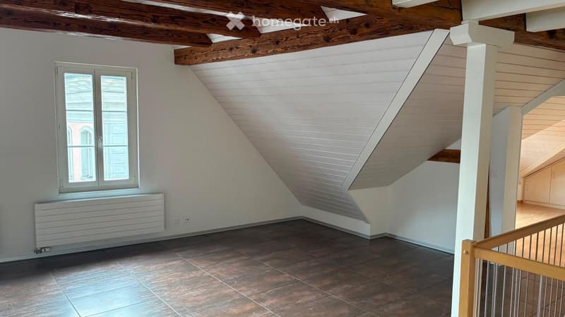 Empty attic room with exposed wooden beams, tiled floor, and a window with white frame. The room features a staircase leading to another area, and a radiator is visible below the window.