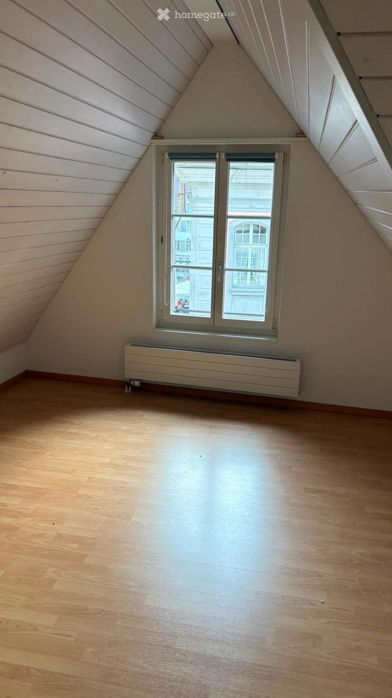 Empty attic room with wooden flooring, white walls, and a single window offering a view of the street.