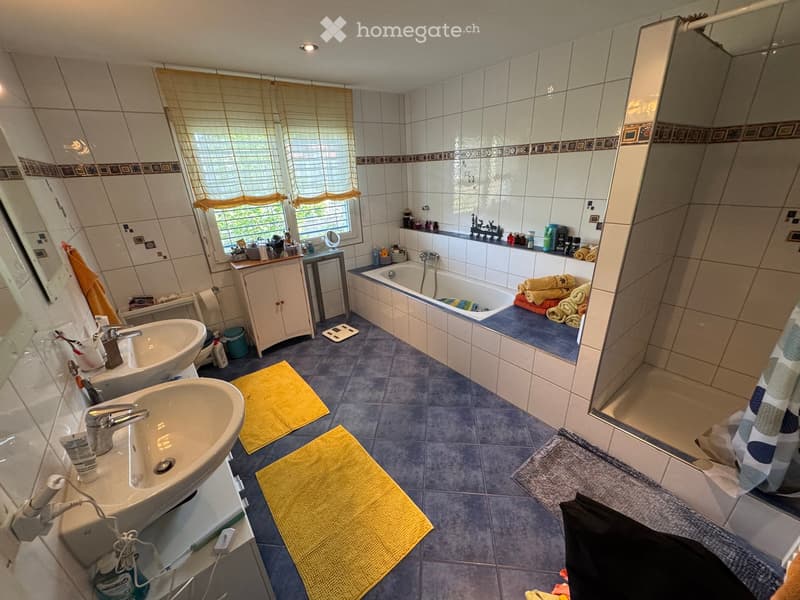 A well-lit bathroom with two sinks, a bathtub, a large mirror, and a shower area. The room features tiled walls and floors, a window with blinds, and various bathroom accessories on the counter.