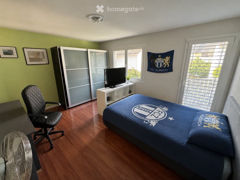 Cozy bedroom with a single bed, a black office chair, a small fan, a wardrobe, a TV on a shelf, and framed pictures on the wall.