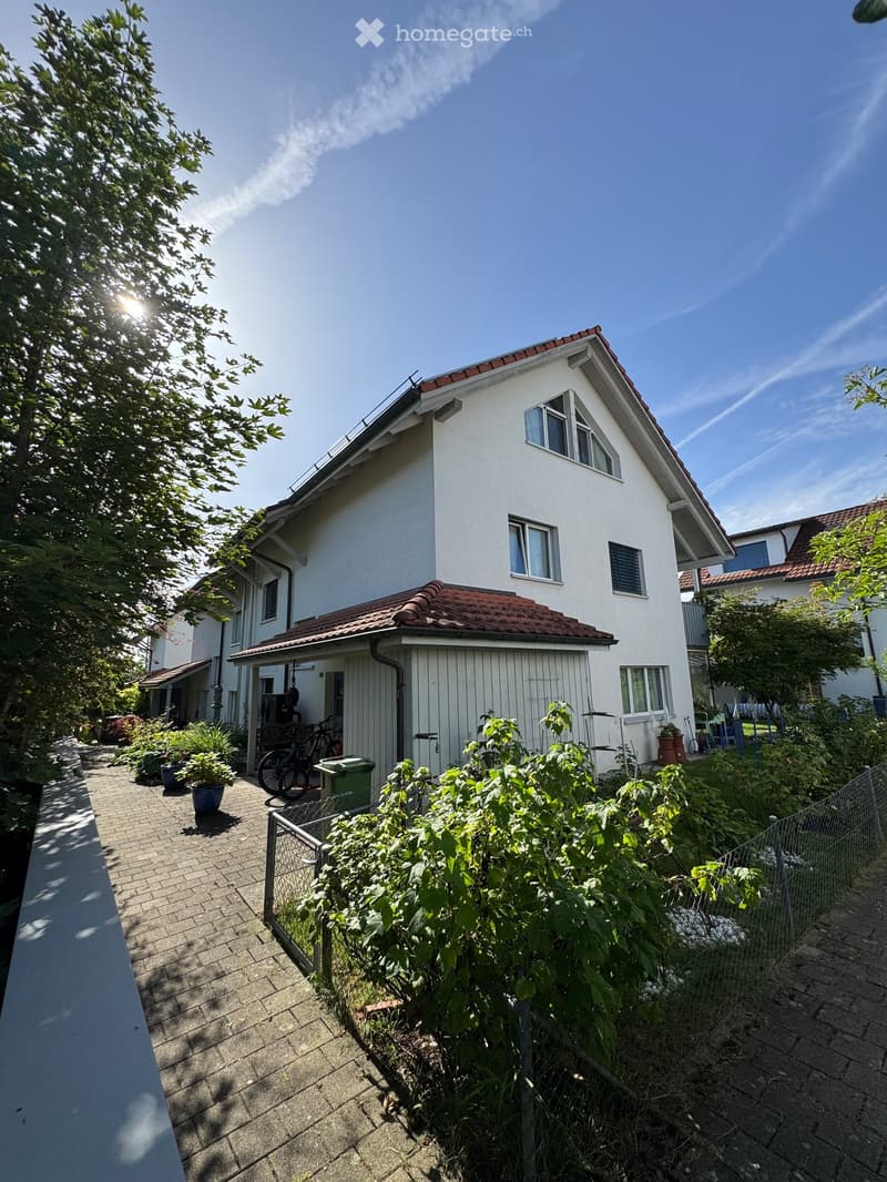 Two-story white house with red-tiled roof, multiple windows, and a small garden area with potted plants and a bicycle.