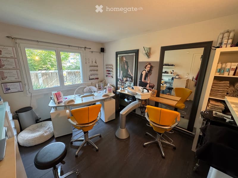 Well-lit salon with two yellow client chairs, a white sink, a large mirror, shelves with nail polish, and a framed picture on the wall.