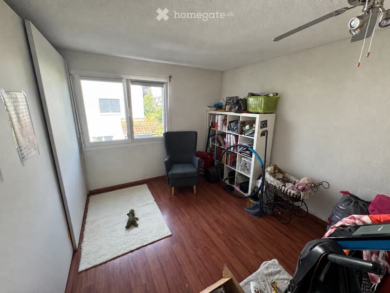 A well-lit room with wooden flooring, a large window, a grey armchair, a bookshelf filled with books and items, a baby stroller, a laptop, and a vacuum cleaner.