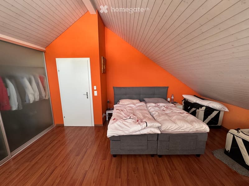 Bright bedroom with wooden flooring, slanted wooden ceiling, and orange walls. Features a double bed with white bedding, a nightstand with a lamp, and a mirrored wardrobe.