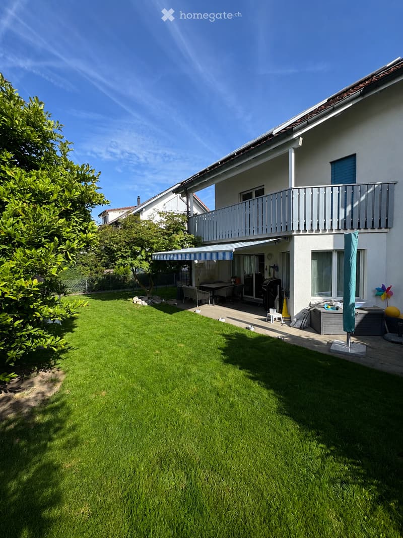 Well-maintained garden with green lawn, patio area with outdoor furniture, barbecue, and a covered seating area. The house features a balcony and large windows.