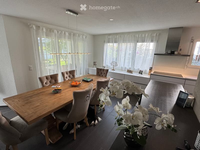Modern dining area with a wooden dining table, six upholstered chairs, a flower vase with white orchids, and a small screen on the floor.