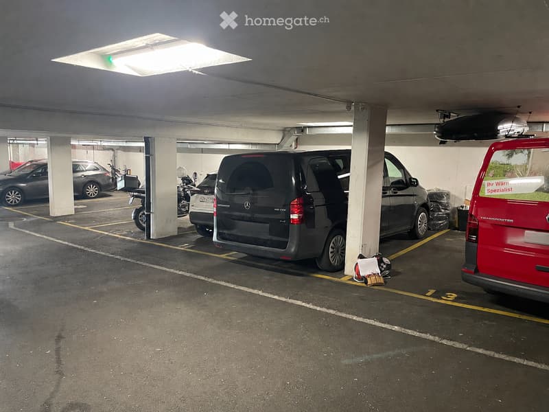 Underground parking area with several parked vehicles, including a Mercedes-Benz van, a red van, and several cars. The area is well-lit with overhead lighting and marked parking spaces.