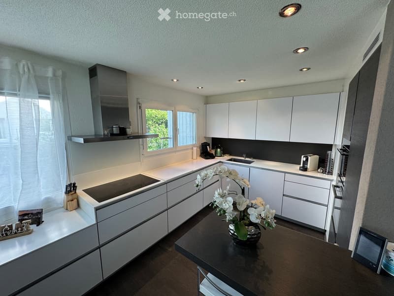 Modern kitchen with white cabinets, induction stove, sink, and various appliances. Features a dark wooden table with a flower vase, and a window with white curtains.