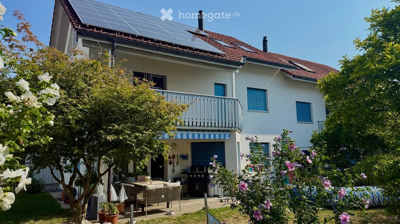 Two-story house with solar panels on the roof, a balcony, and a well-maintained garden with outdoor seating and a barbecue.