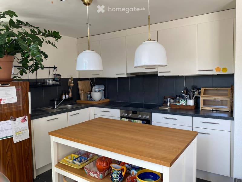 Modern kitchen with white cabinets, dark countertops, and a wooden island. Features pendant lights, a sink, oven, and various kitchen appliances and utensils.