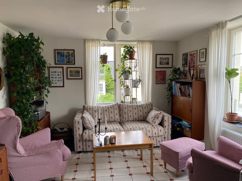 Living room with patterned sofa, coffee table, armchairs, bookshelf, and decorative plants.
