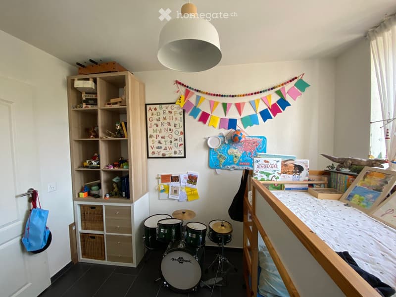 A child's room with a drum set, a wooden shelf filled with books and toys, a world map, an alphabet poster, and a bunk bed with a framed picture on the wall.