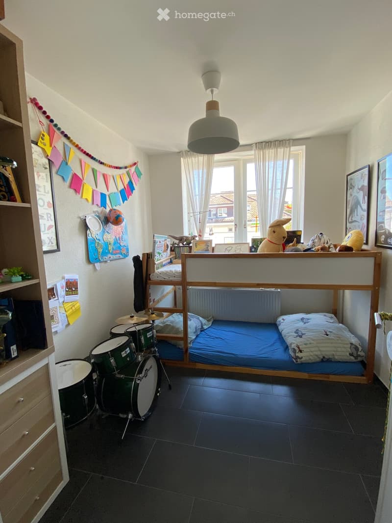 A cozy bedroom with a bunk bed, a drum set, a shelf with books and toys, and colorful decorations.