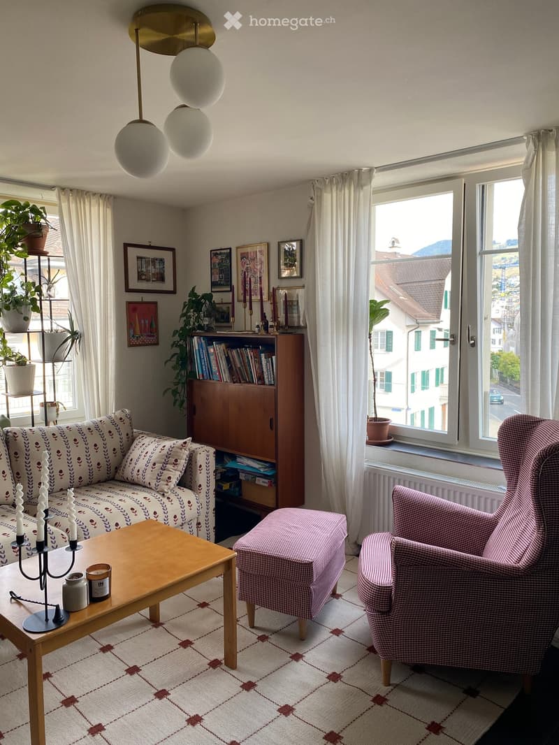 Cozy living room with a patterned sofa, wooden coffee table, armchairs, bookshelf, and decorative items. Large windows offer a view of the city.