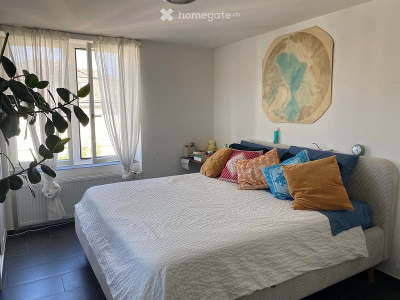 A cozy bedroom with a neatly made bed, colorful pillows, a nightstand with a clock, and a decorative wall piece. The room features large windows with white curtains and a plant in the corner.