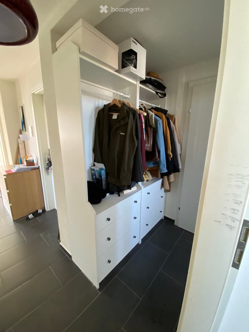 A well-lit hallway with a built-in white wardrobe unit featuring multiple drawers and hanging space for coats and jackets. The floor is dark tiled, and the walls are light-colored.