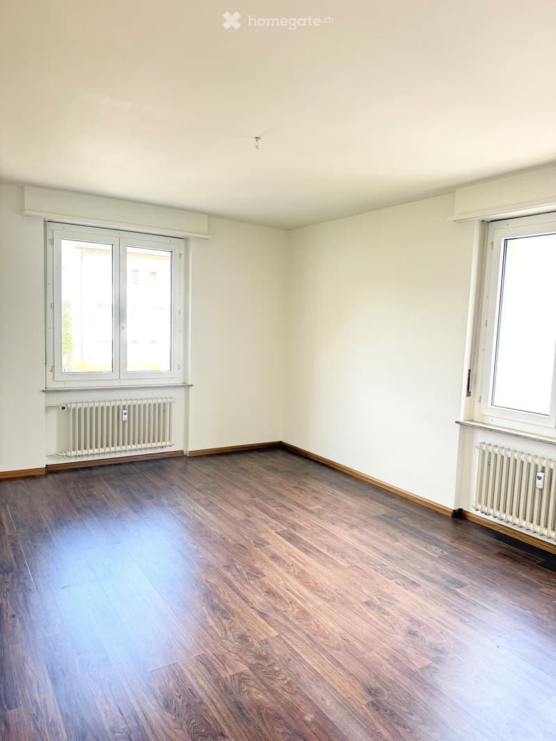 Empty room with wooden flooring, white walls, and large windows with radiators beneath them.