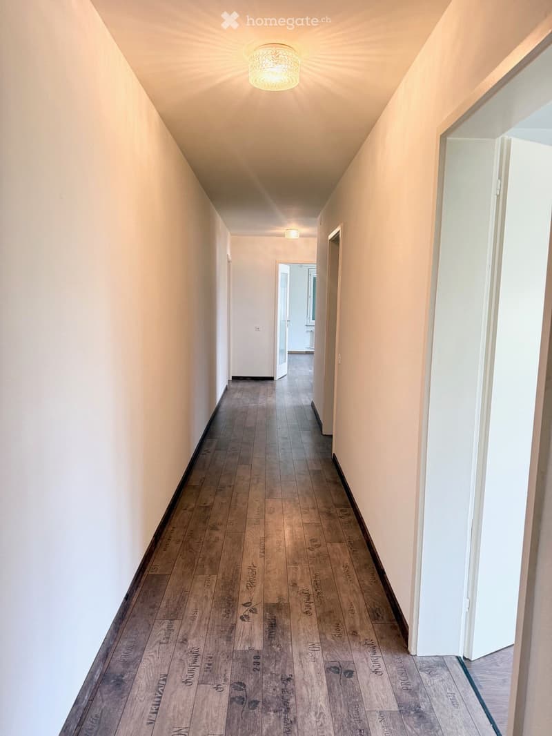 A long, narrow hallway with wooden flooring and white walls, featuring recessed lighting and several doors leading to other rooms.