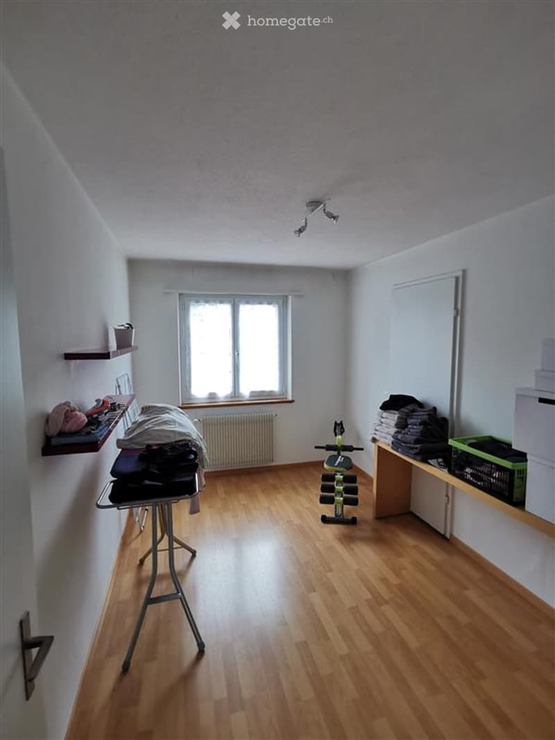 Furnished Room. The room features wooden flooring, white walls, and a large window allowing natural light. There is a table with folded clothes, shelves with various items, an exercise bike, and weights in the corner.