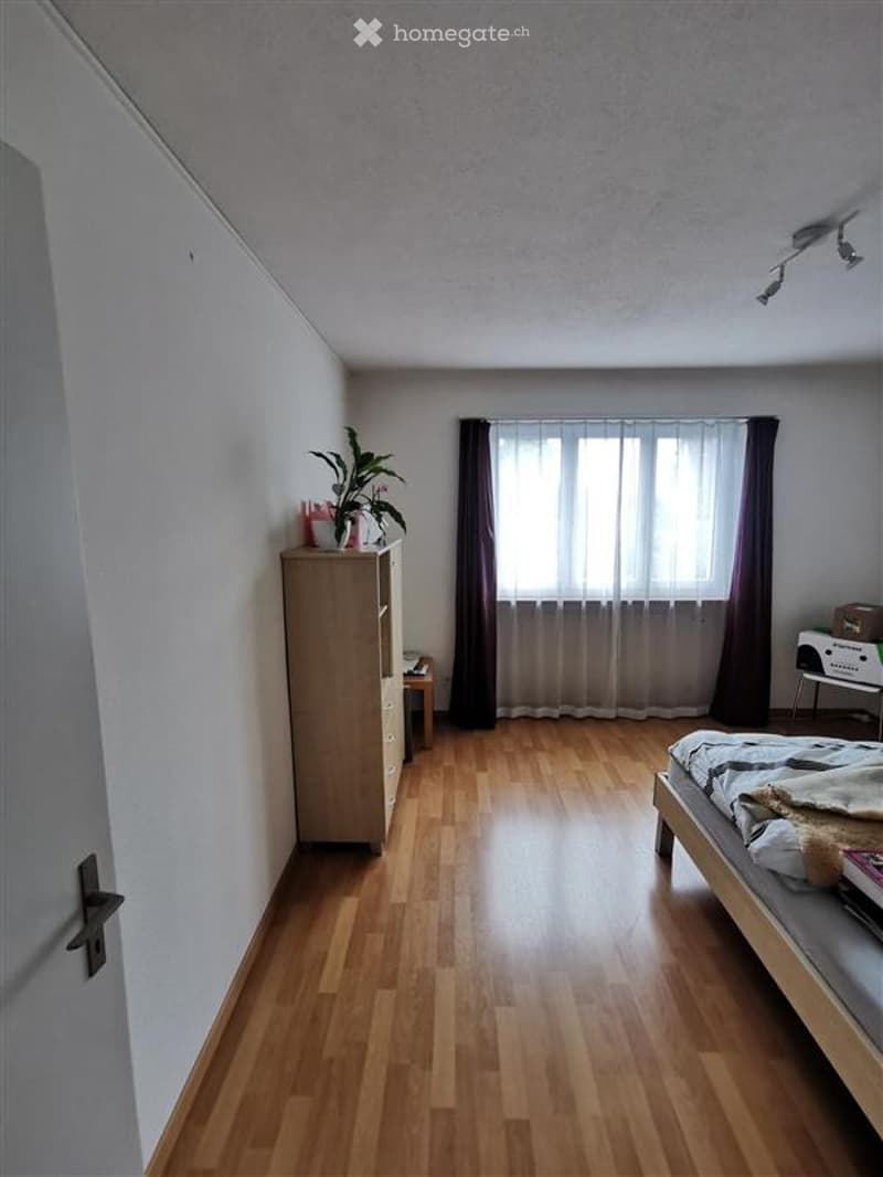 Bedroom. A well-lit bedroom with wooden flooring, a bed with a patterned duvet, a wardrobe, a small table with a plant, and a window with curtains.