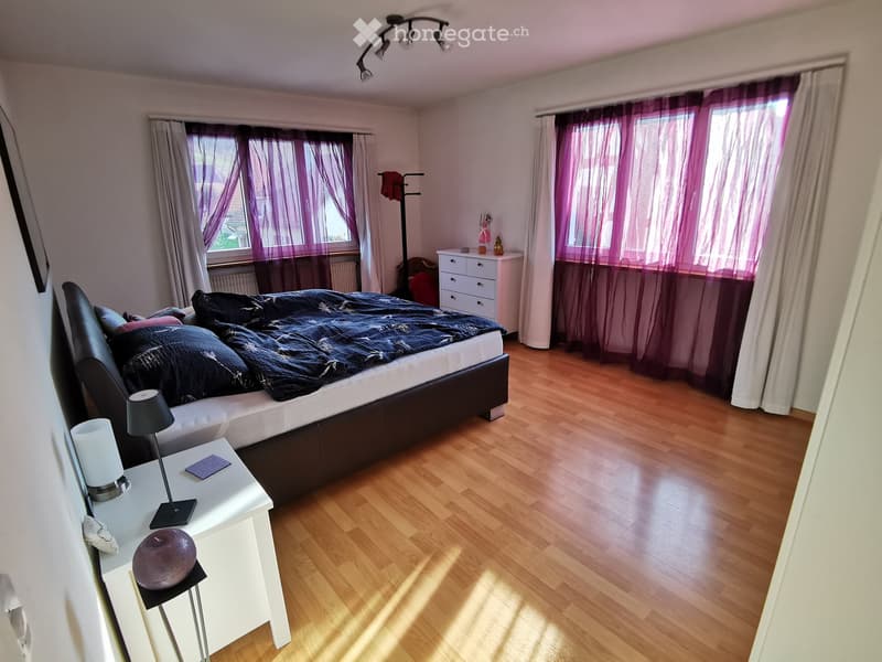 Bedroom. Well-lit bedroom with wooden flooring, large windows with purple curtains, a black leather bed, nightstand with lamp, and a small drawer unit.