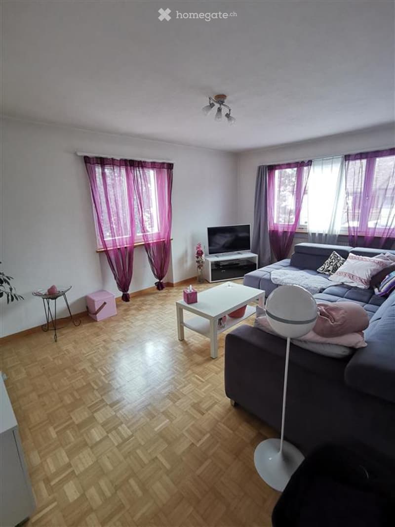 Living Room. Modern living room with wooden flooring, large windows with purple curtains, grey sectional sofa, white coffee table, TV stand, and decorative items.