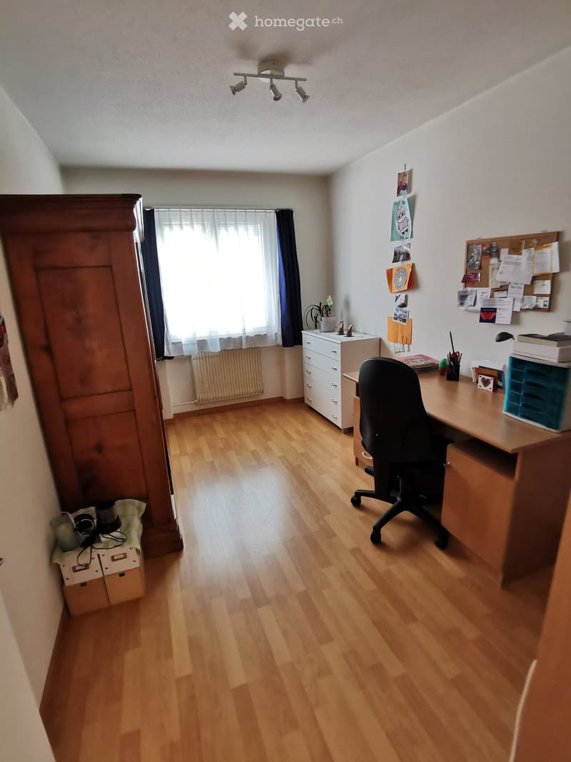 Furnished Room. A well-lit study or home office with wooden flooring, a large window with curtains, a desk with a chair, a cabinet, a drawer unit, and various office supplies.