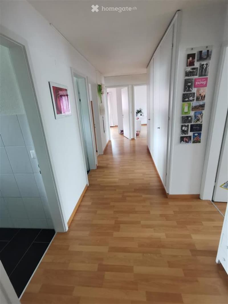 Corridor / Hallway. A long, well-lit hallway with wooden flooring and white walls. Doors and framed pictures are visible along the walls, leading to other rooms.