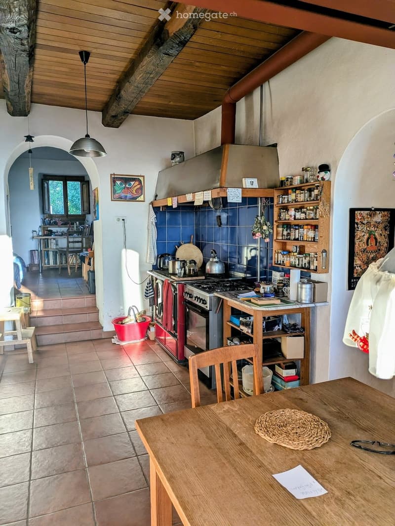 Cozy kitchen with wooden beams, tiled floor, and blue tiled backsplash. Includes a wooden dining table, chairs, stove, oven, and shelves with various items.