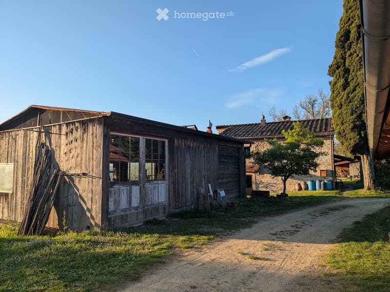 A rustic wooden shed with a slanted roof and large windows, situated on a grassy area with a dirt path leading to it. In the background, there is a stone building with a tiled roof and several blue barrels.