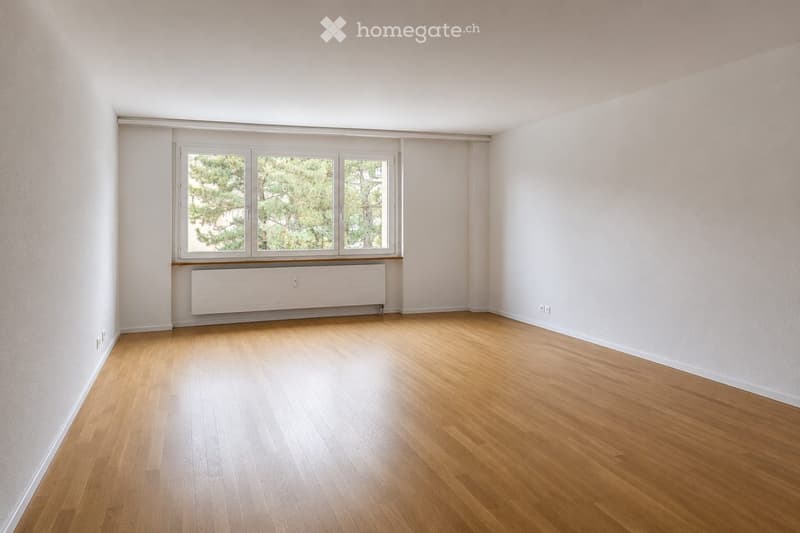 Empty room with light wooden flooring, white walls, and large windows with a radiator below.