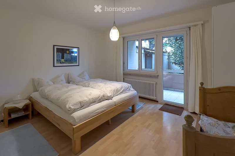 Cozy bedroom with a wooden bed, nightstand, and large windows leading to a balcony. The room features light wooden flooring and a picture frame on the wall.