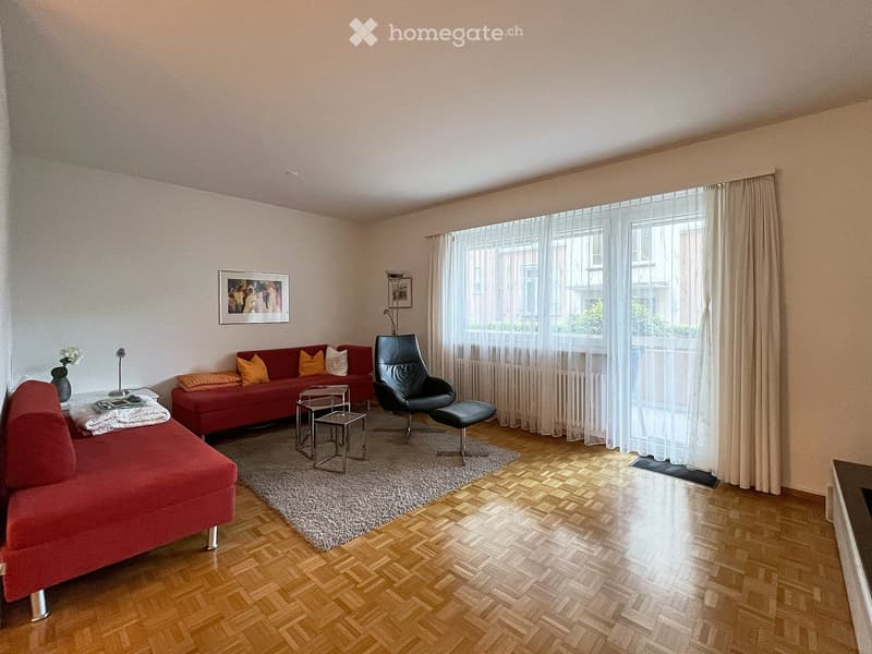 Modern living room with wooden flooring, red sofa, black leather chair, small tables, and decorative items.