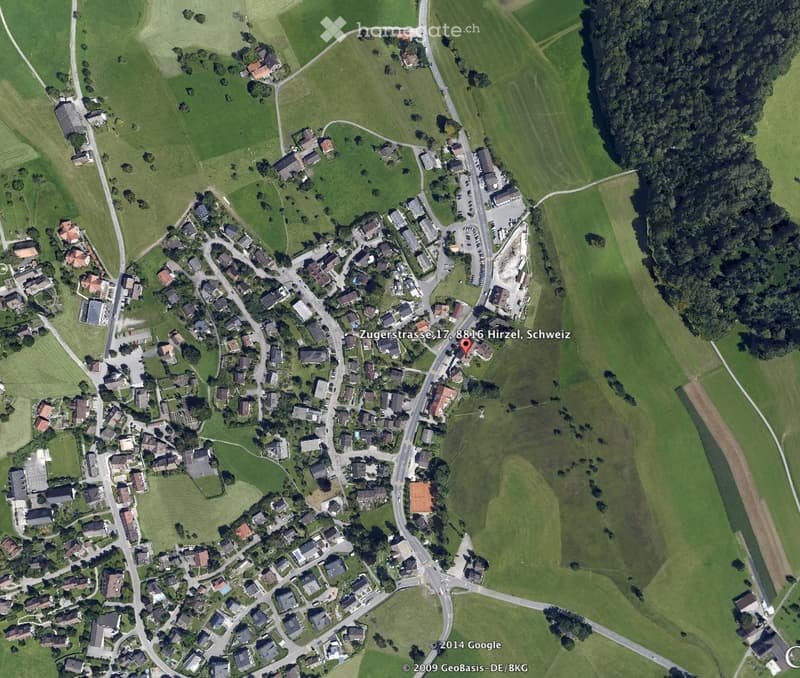 Aerial view of a rural village with scattered houses, green fields, and roads. The area includes residential buildings, agricultural land, and a small cluster of structures near a road intersection.
