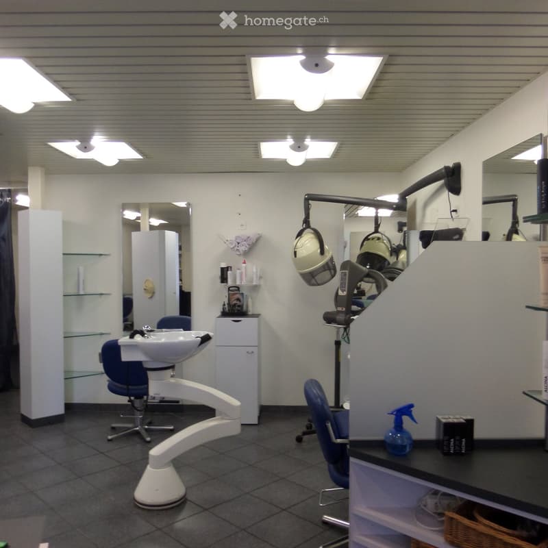 Modern salon with white walls, tiled floor, multiple chairs, sinks, mirrors, hair dryers, and shelves holding various products.