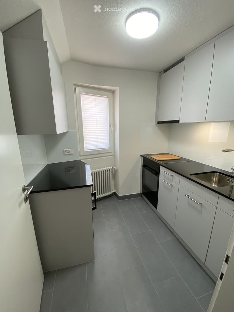 Modern kitchen with white cabinets, dark countertops, and grey tiled flooring. Features a sink, oven, and a small window with blinds.