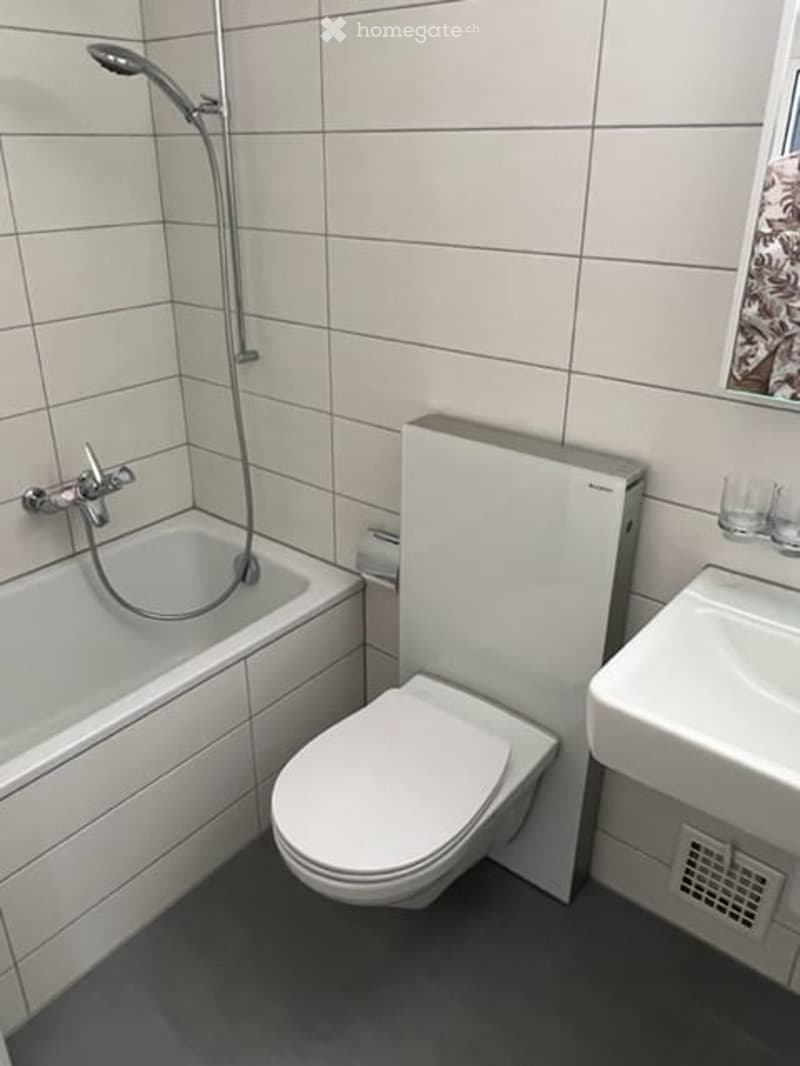 Modern bathroom with a white bathtub, toilet, sink, and showerhead. The walls are tiled in light gray, and the floor is dark gray.