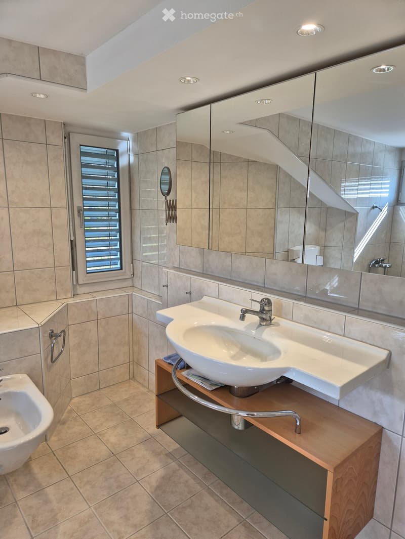 Modern bathroom with a white sink, wooden vanity, large mirror, and a bidet. Features tiled walls and floor, recessed lighting, and a window with blinds.