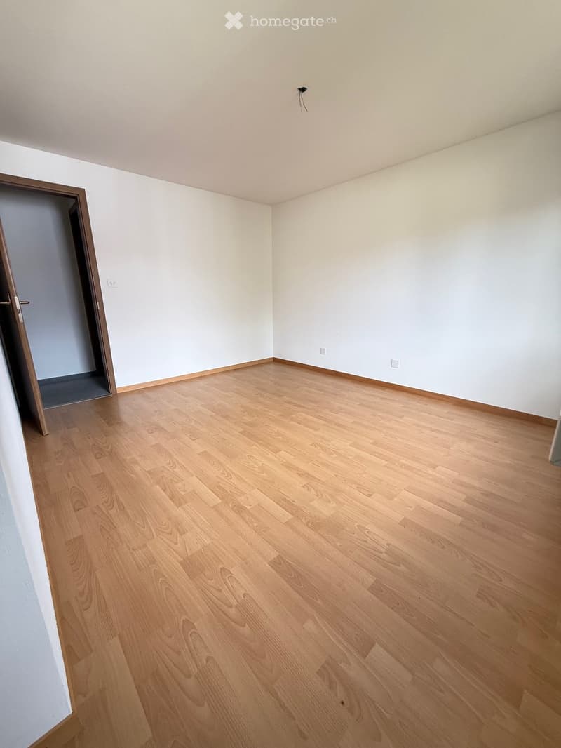 Empty room with light wooden flooring and white walls, featuring a doorway on the left side.