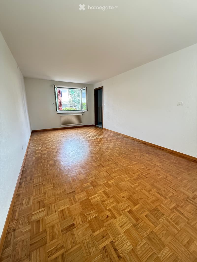Empty room with wooden herringbone flooring, white walls, and a window with a radiator below it. The room has a doorway leading to another space.