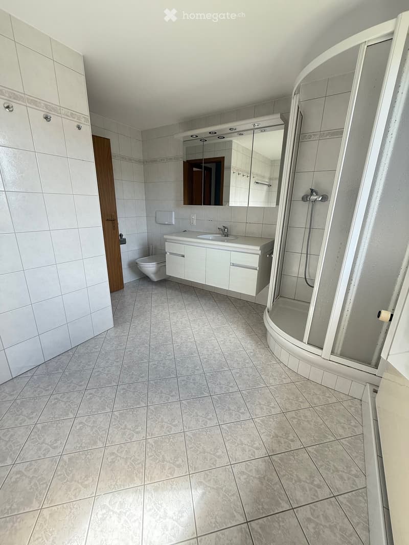Modern bathroom with white tiles, a sink, toilet, mirror, and a glass-enclosed shower. The space is clean and well-lit with a wooden door visible on the left side.